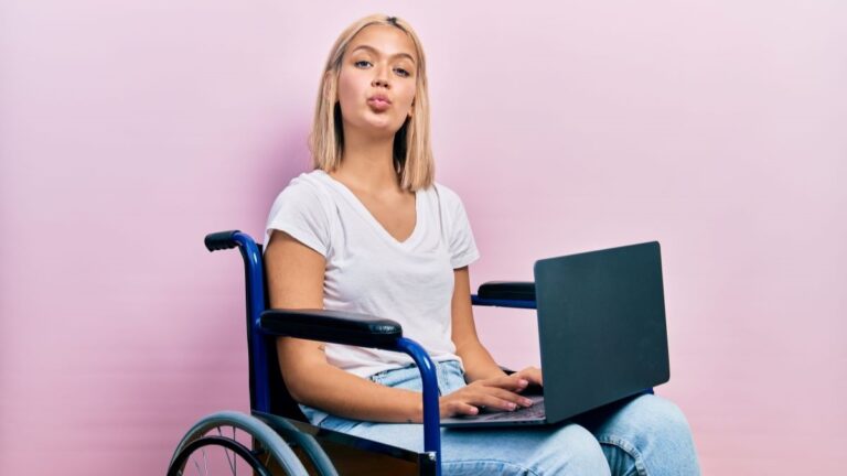 Cheeky young blonde woman in a wheelchair puckers her lips while working on a laptop