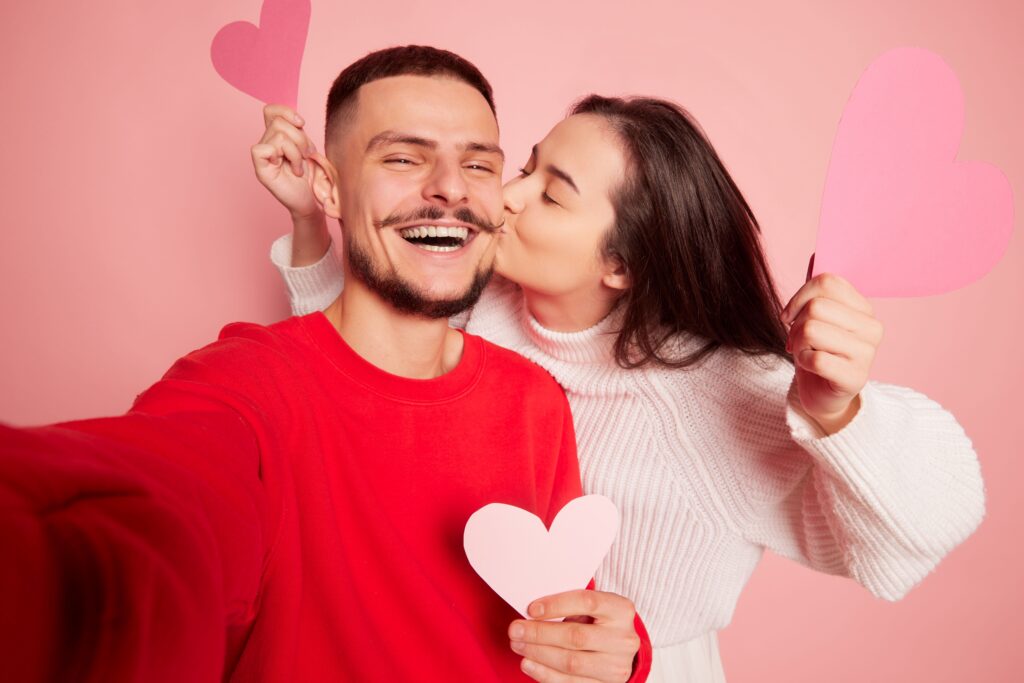 white-young-couple-woman-kissing-cheek-both-holding-paper-hearts-against-a-pink-background