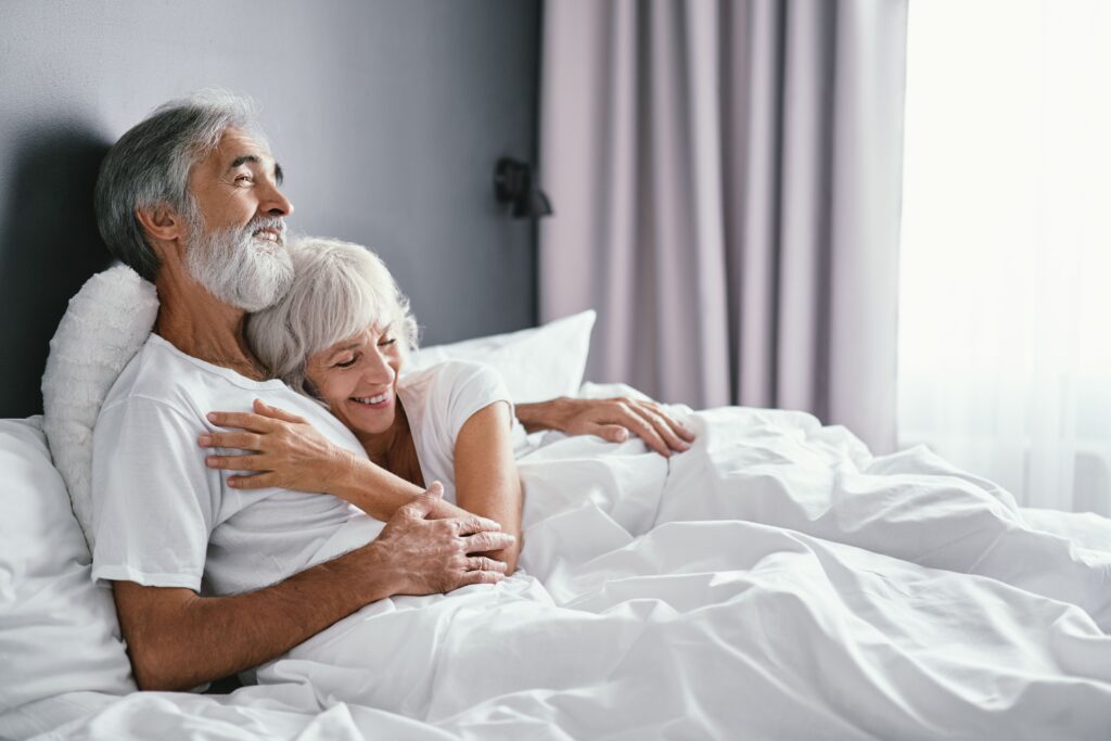 An older couple, both with grey hair, laying in bed snuggling. This is showing how dementia affects sexual relationships over time. 