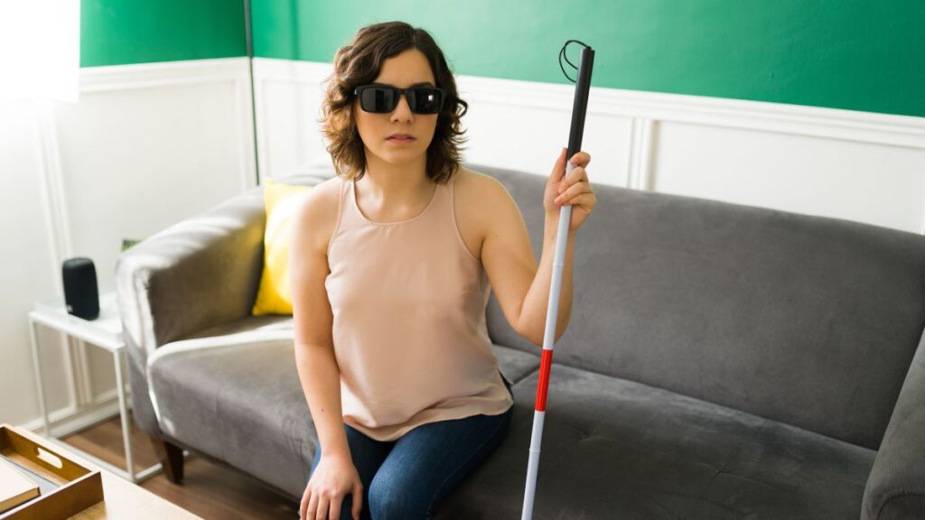 young white blind woman wearing dark sunglasses sitting on a coach and holding a white cane