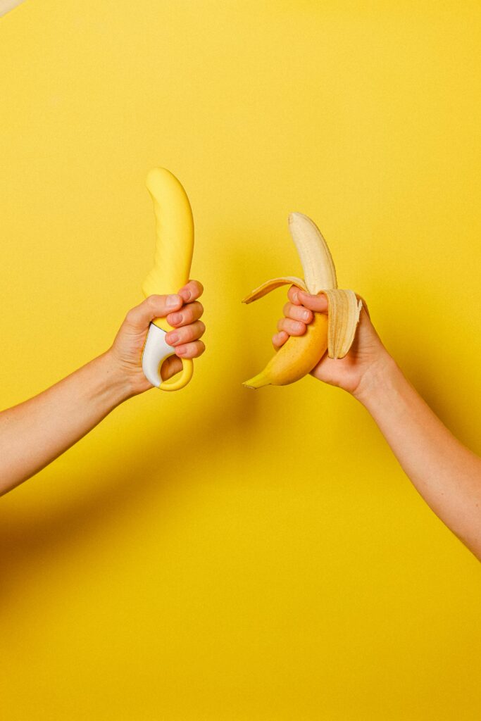 A hand holding a yellow vibrator next to a hand holding a yellow banana against a yellow background. 