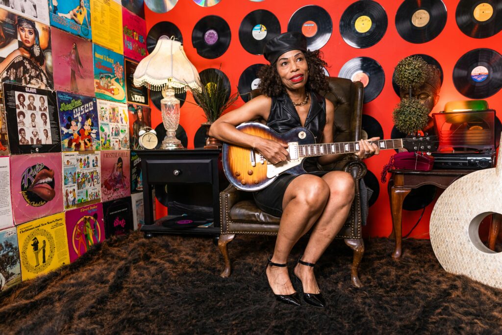 An older black woman sitting in a recording studio surrounded by albums covers and vinyl's. She is playing a guitar. 