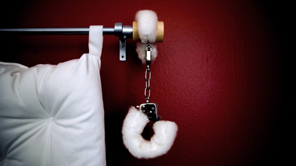 white handfcuffs attached to white bed posts before red wall