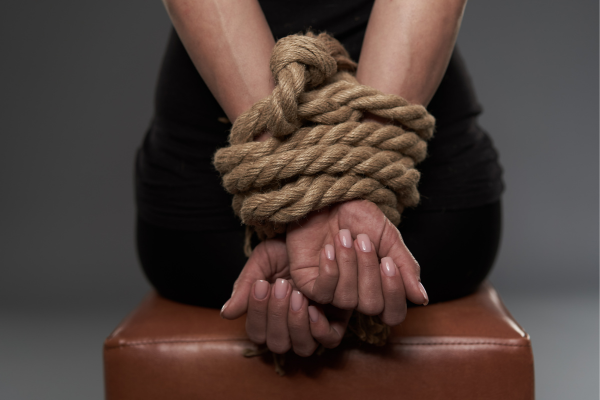 Female sitting on a bench with her hands behind her, bound in thick ropes. 