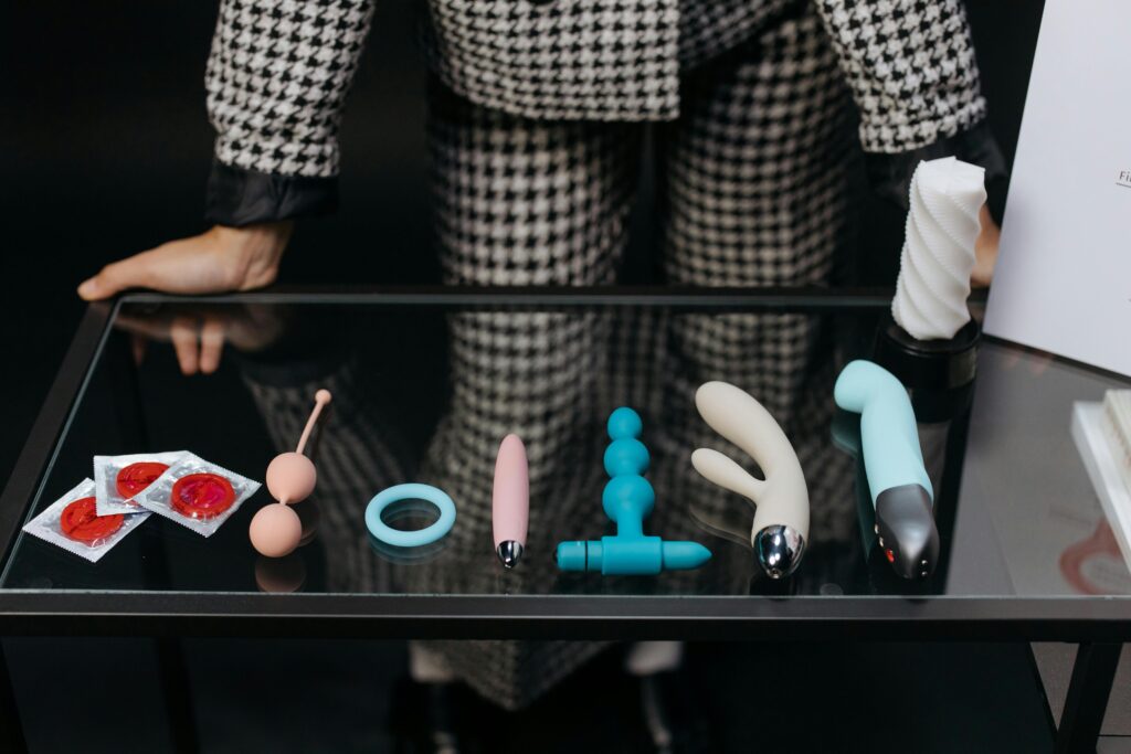 An assortment of sex toys: condoms, kegel balls, cock rings, vibrators, and dildos all sitting on a glass table. With a female in checkered suit standing behind them. 