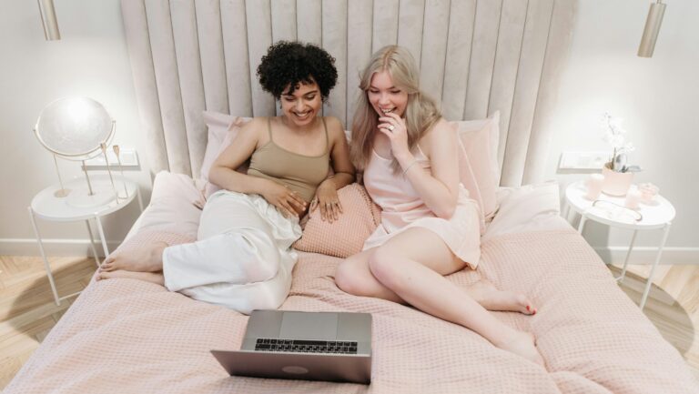 two women lay in bed watching a laptop together
