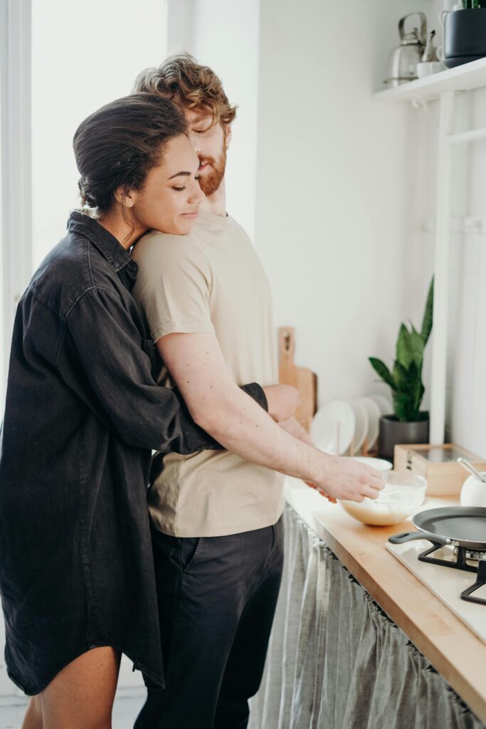 couple snuggling in the kitchen. The women stands behind the man and holds his waist as he mixes food in a bowl. 