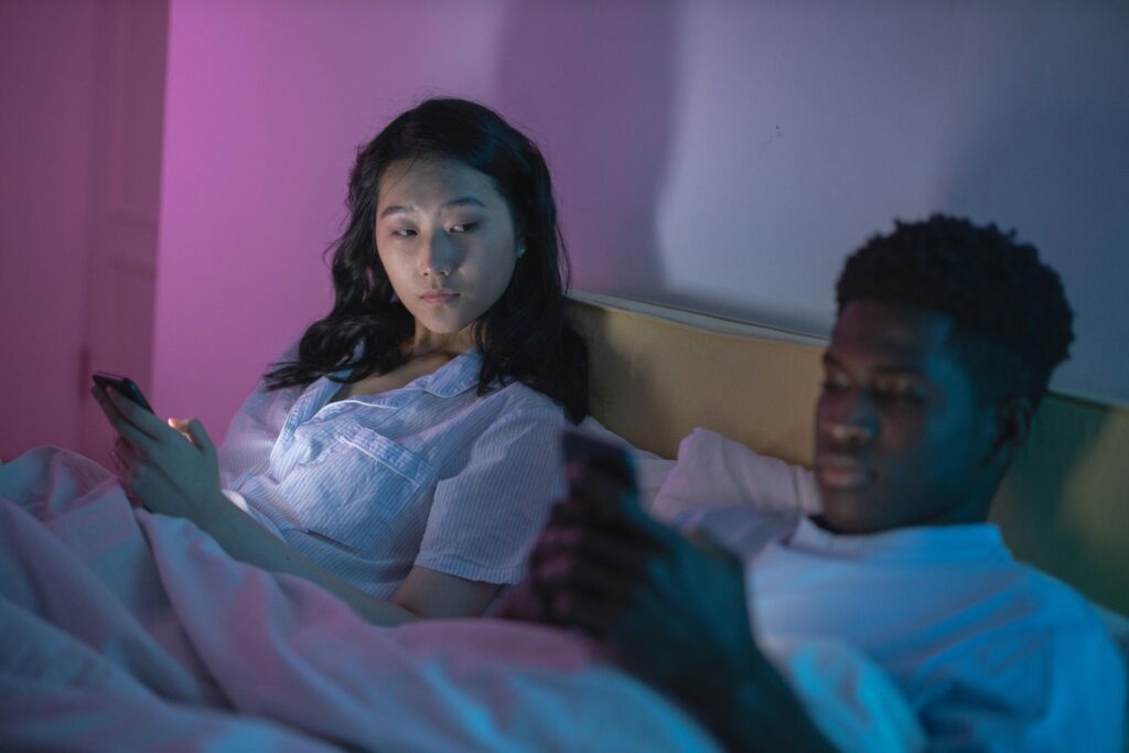 A diverse couple laying in bed on their phones. The female partner is looking at the male partner suspiciously. 