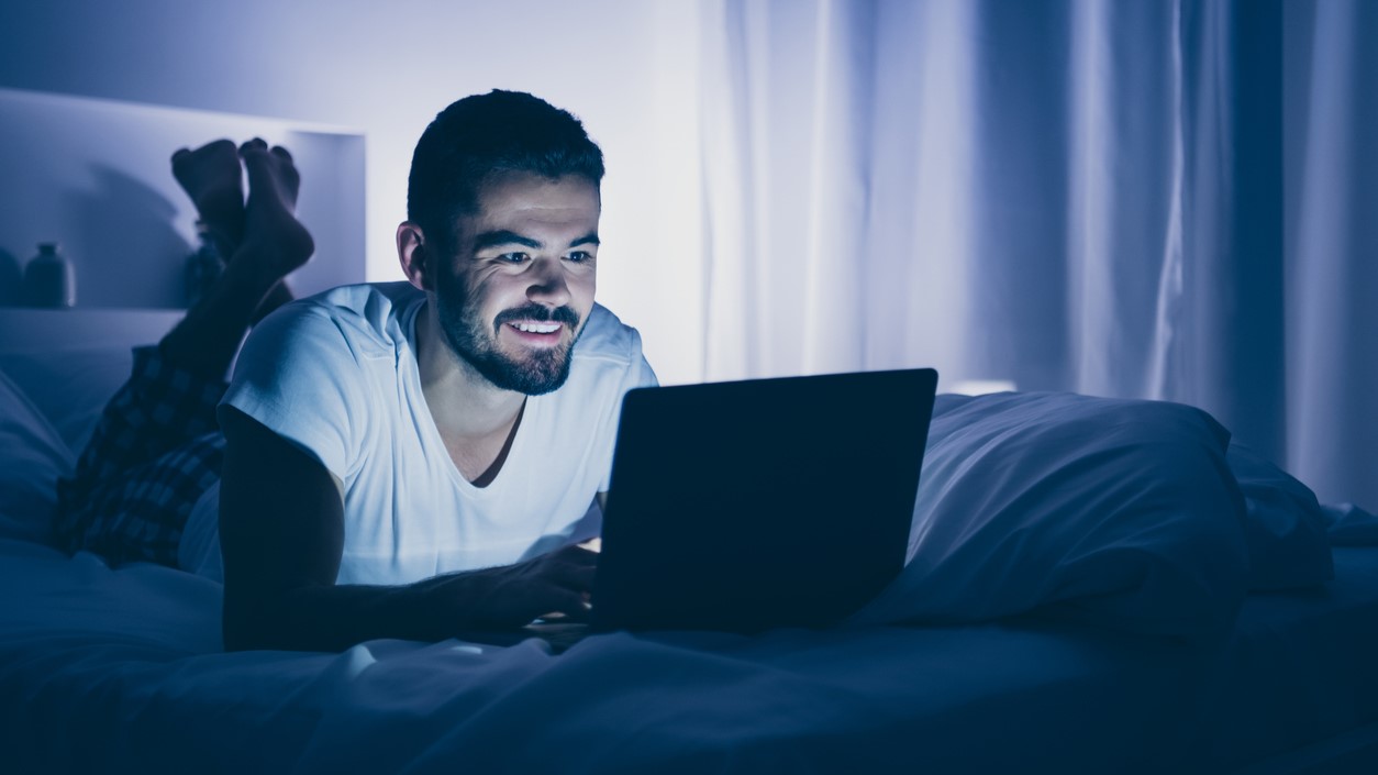 Young man in bed on stomach looking at computer in dark room