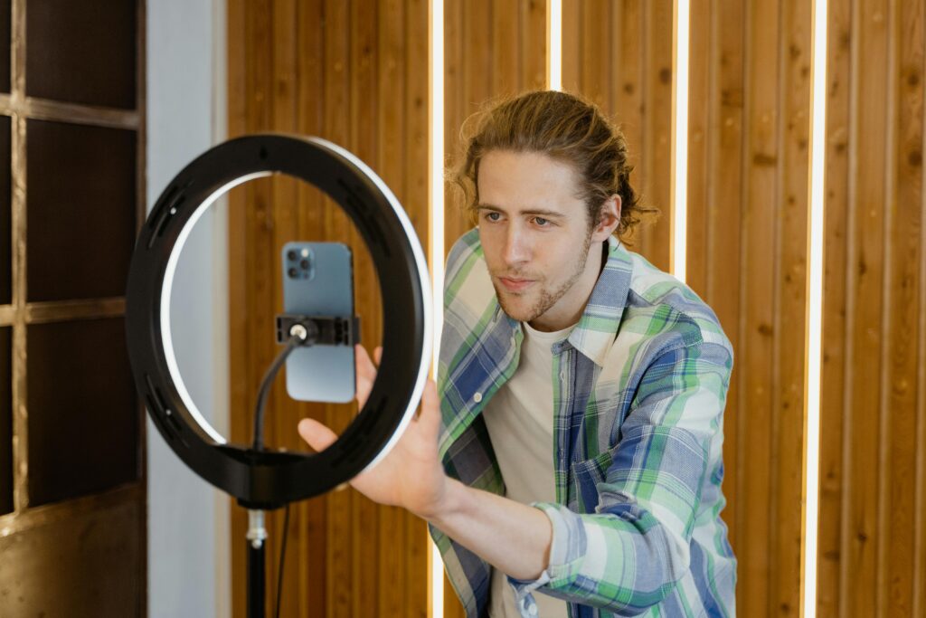 A young male with long hair in a bun stands in front his phone, which is connected to a stand with a ring light. He is adjusting something on his phone and has a concentrated look. 