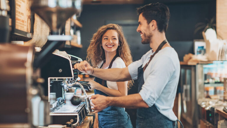 Couple of baristas working in a coffee shop smiling at each other in a flirty way.