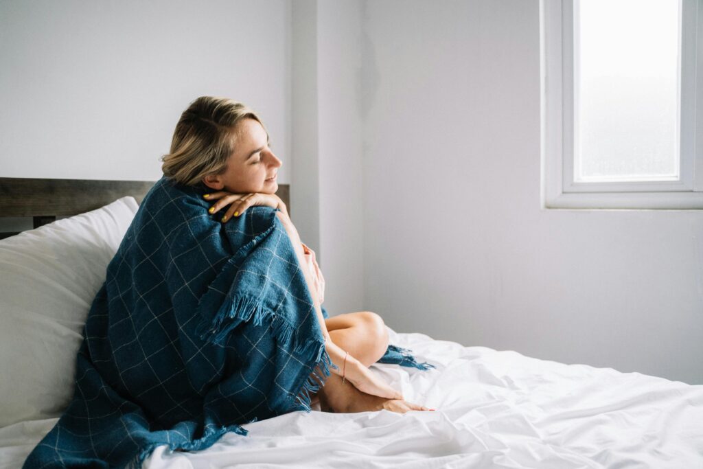 young woman sits on bed, snuggled in a blanket, looking relaxed and happy.