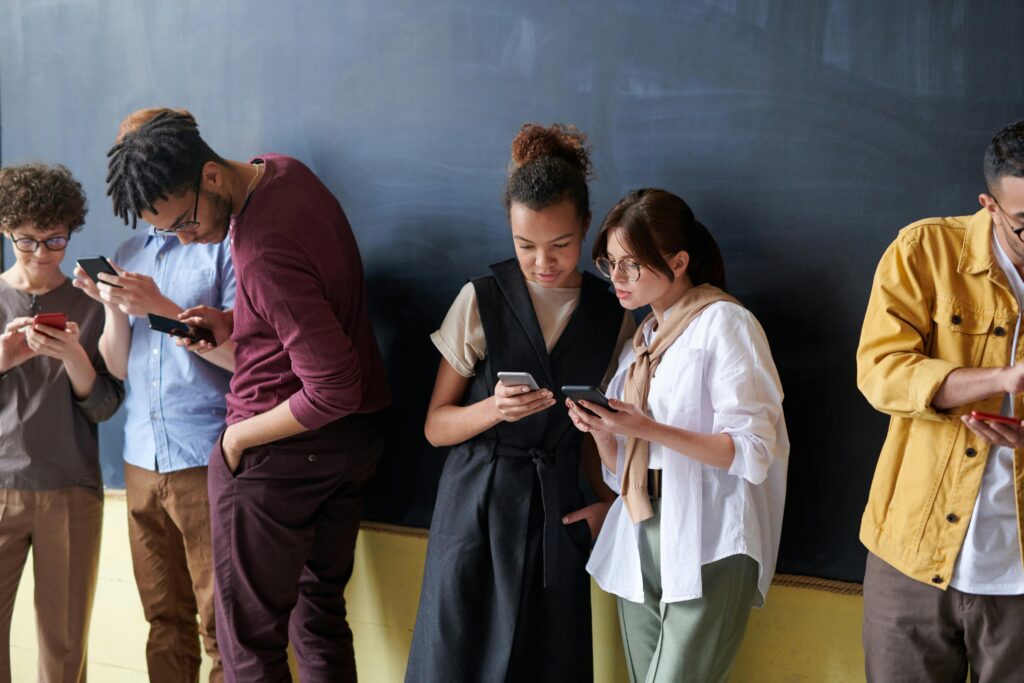 A group of young adults standing in a classroom. They are separated into groups, looking at cellphones. This could be a tech-savvy sex education class. 