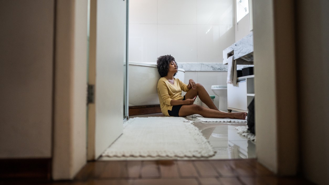 Sad young woman holding a pregnancy test sitting on ground in the bathroom at home